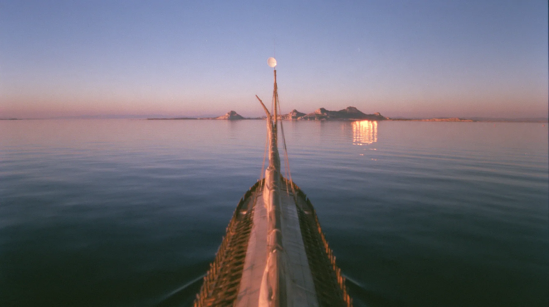 low angle up the mast of a long black ancient Greek galley with a single mast, a square sail and two banks of oars, a small white satellite dish at the top, distant rocks glowing gold on the horizon, dusk, low gold…
