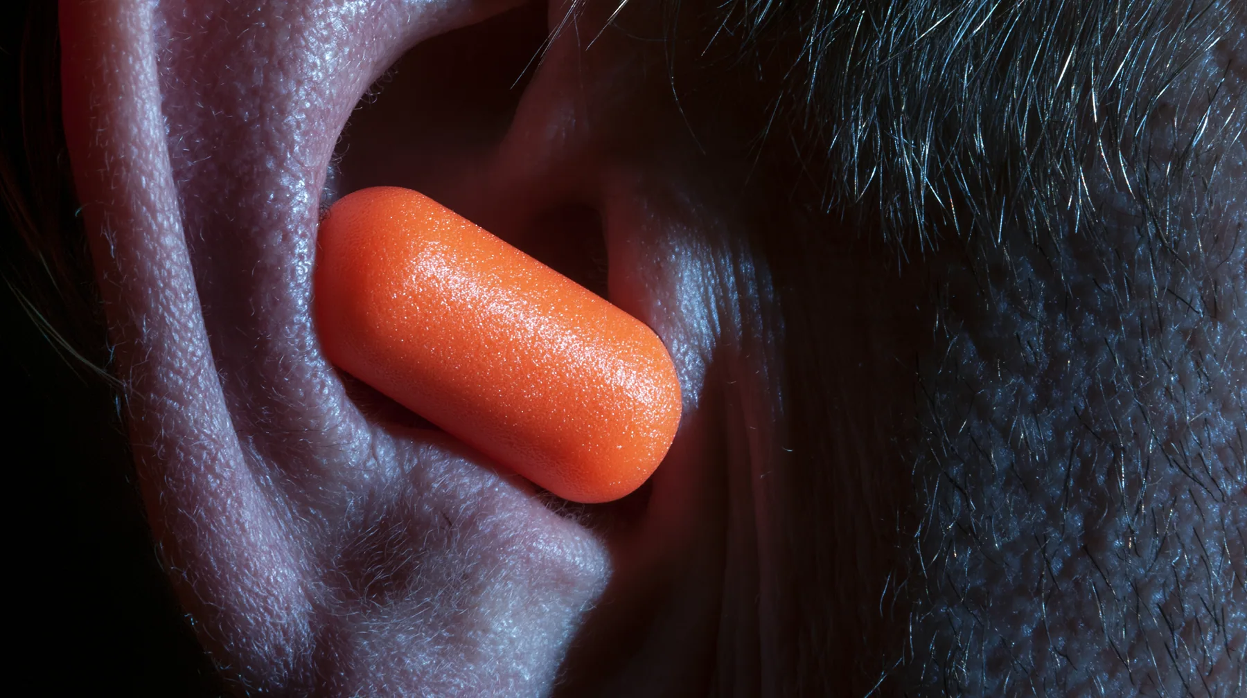 macro photograph of a bright orange foam earplug expanding inside a man's ear, macro product photograph, hard rim light, razor-thin focus, dark background, no text, no letters, no logos