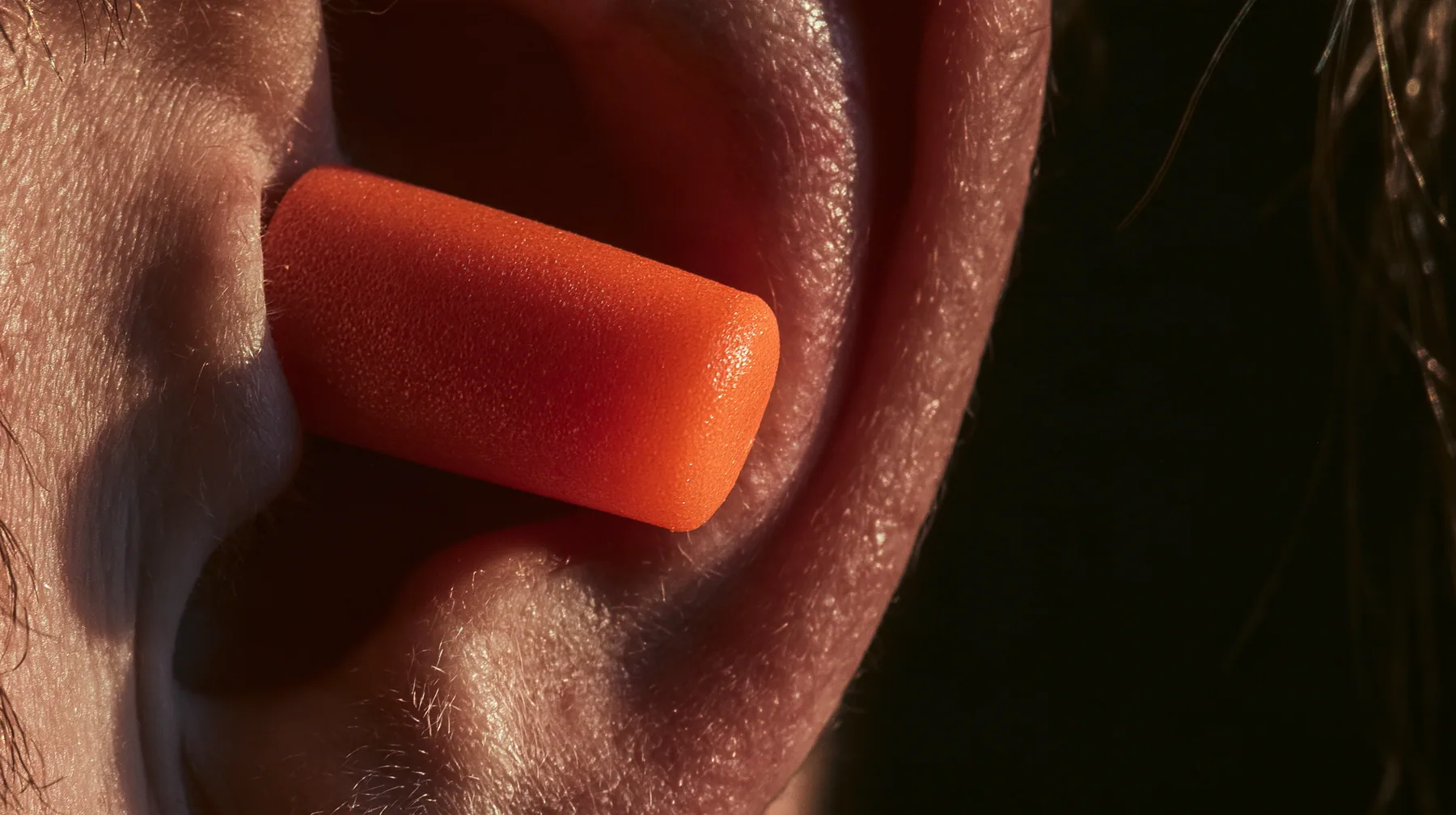 macro photograph of a bright orange foam earplug expanding inside a man's ear, macro product photograph, hard rim light, razor-thin focus, dark background, no text, no letters, no logos