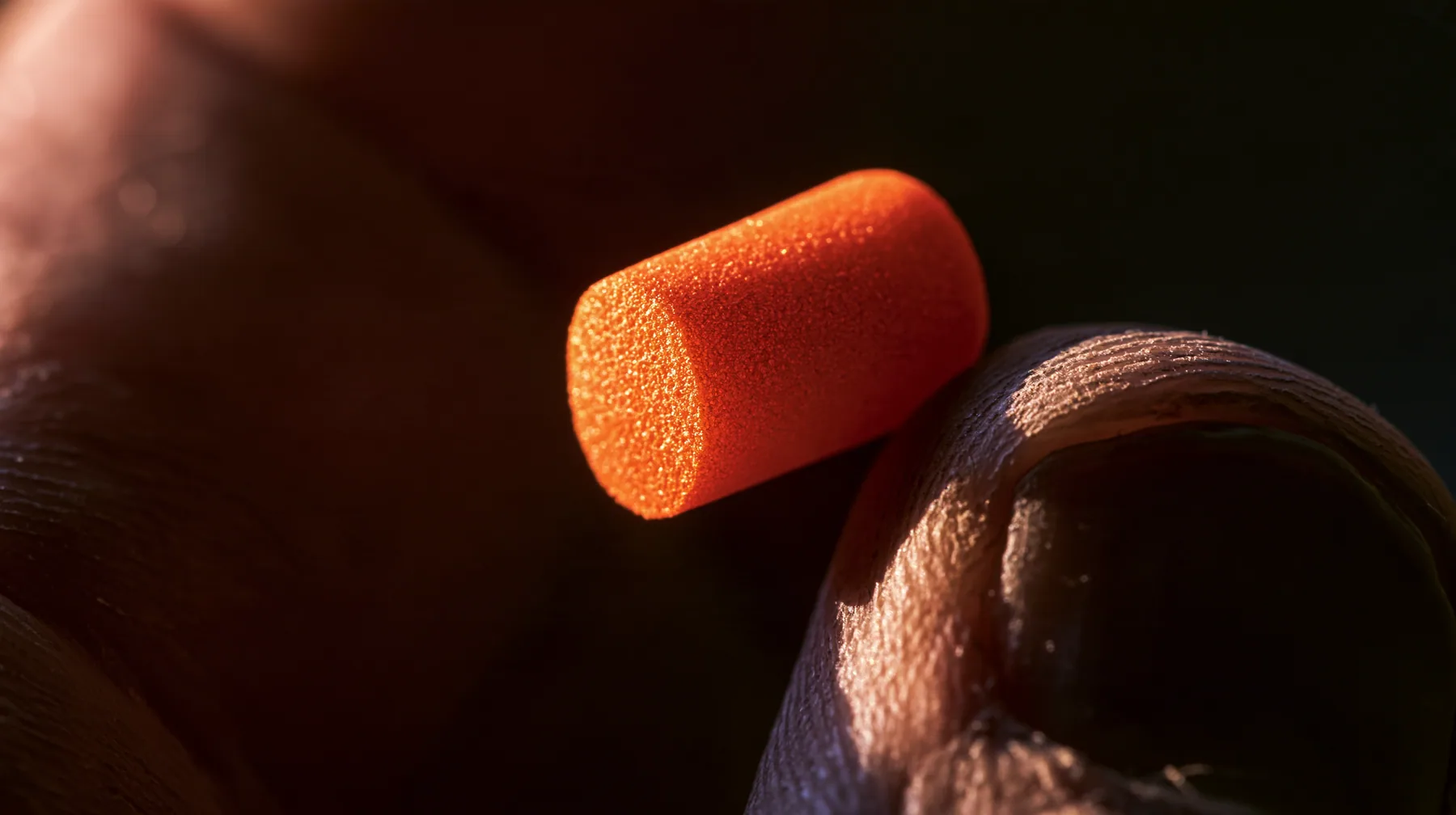 macro of a rower's rough fingers rolling a bright orange foam earplug, macro product photograph, hard rim light, razor-thin focus, dusk light, no text, no letters, no logos
