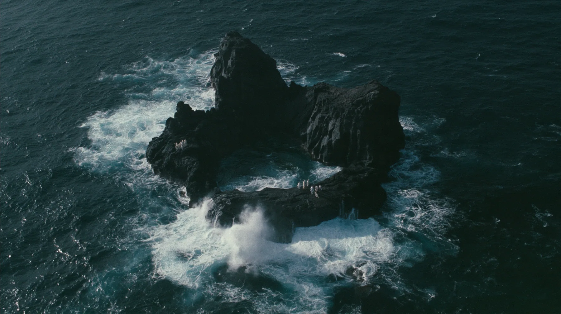 aerial photograph looking straight down at a ring of jagged black volcanic sea stacks in a dark sea, surf glowing white around them, tiny pale figures standing on the rocks, night, moonlight on a black-teal sea,…