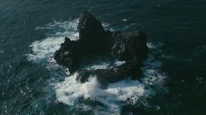 aerial photograph looking straight down at a ring of jagged black volcanic sea stacks in a dark sea, surf glowing white around them, tiny pale figures standing on the rocks, night, moonlight on a black-teal sea,…