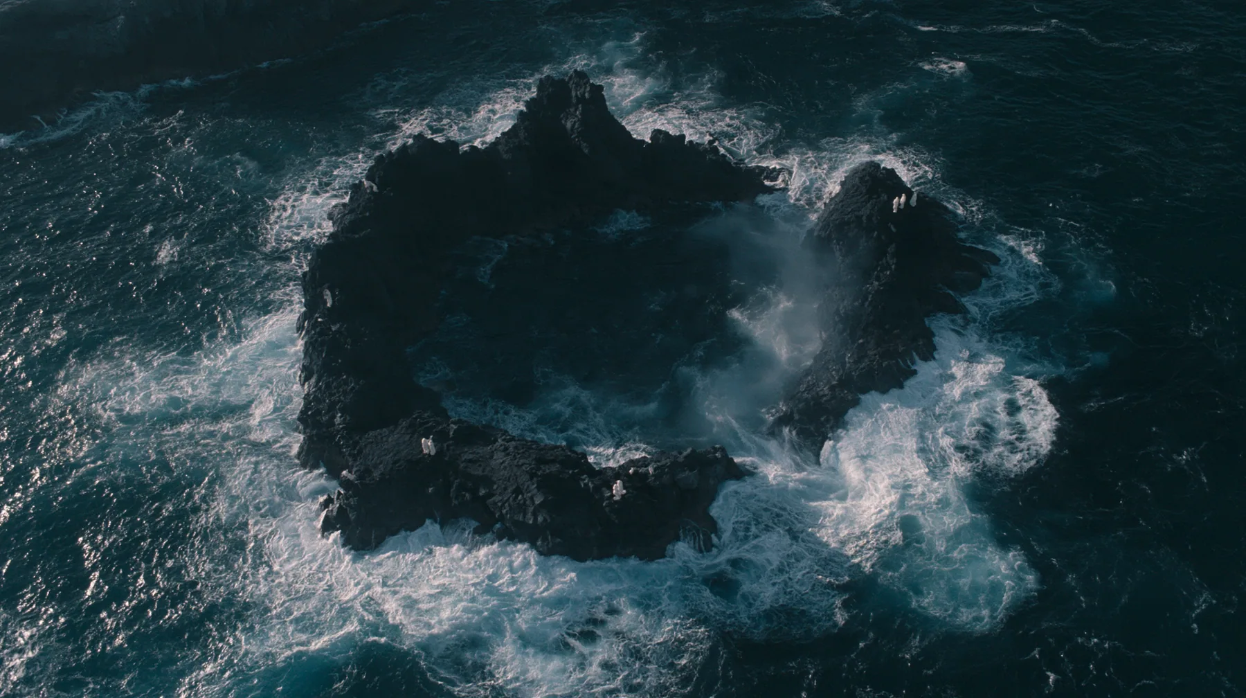 aerial photograph looking straight down at a ring of jagged black volcanic sea stacks in a dark sea, surf glowing white around them, tiny pale figures standing on the rocks, night, moonlight on a black-teal sea,…