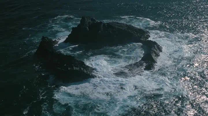 aerial photograph looking straight down at a ring of jagged black volcanic sea stacks in a dark sea, surf glowing white around them, tiny pale figures standing on the rocks, night, moonlight on a black-teal sea,…