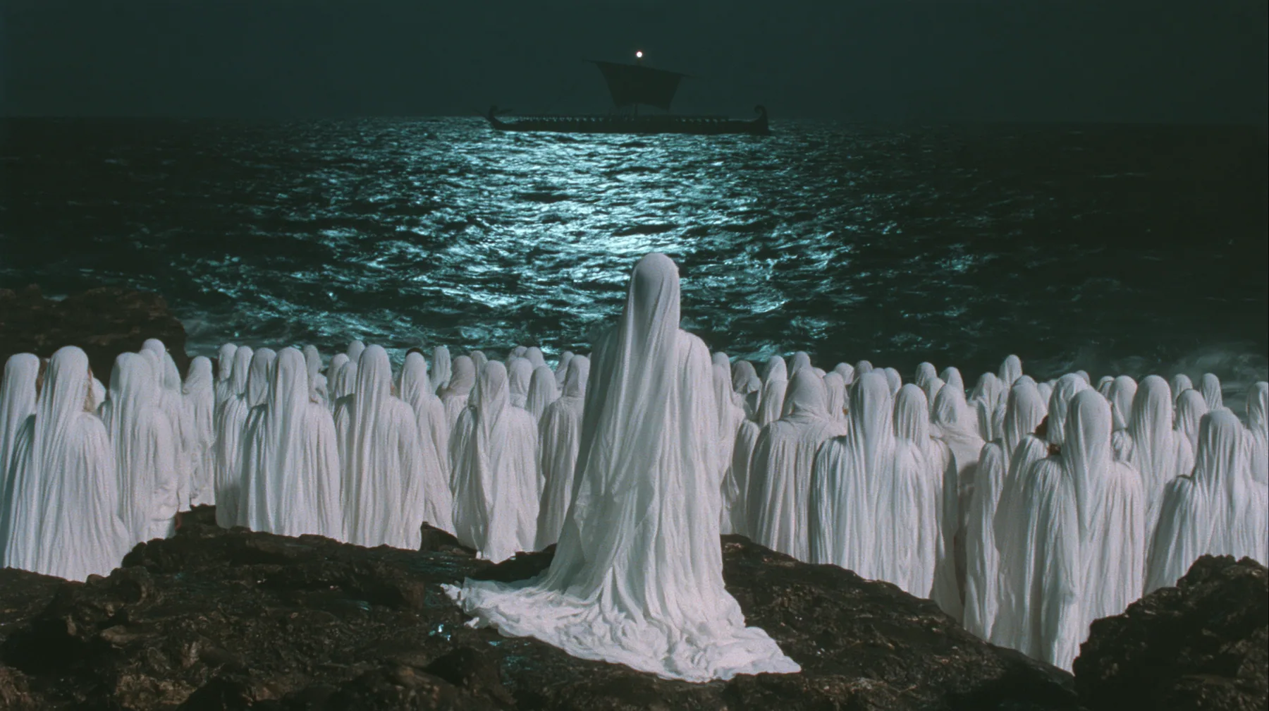 extreme wide shot from behind hundreds of women in white gowns on black sea rocks, looking out to a long black ancient Greek galley with a single mast, a square sail and two banks of oars on the far horizon, one…