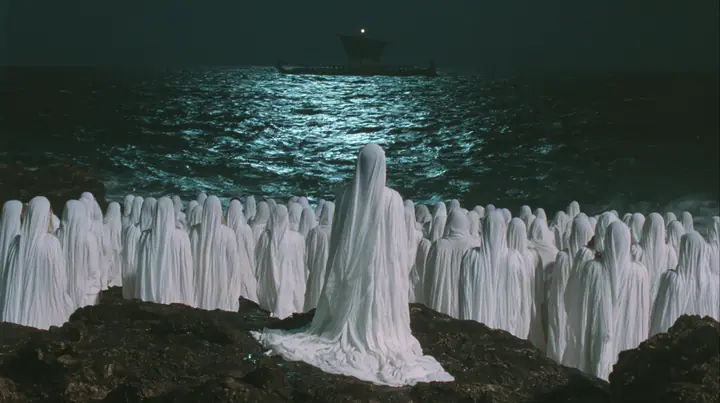 extreme wide shot from behind hundreds of women in white gowns on black sea rocks, looking out to a long black ancient Greek galley with a single mast, a square sail and two banks of oars on the far horizon, one…