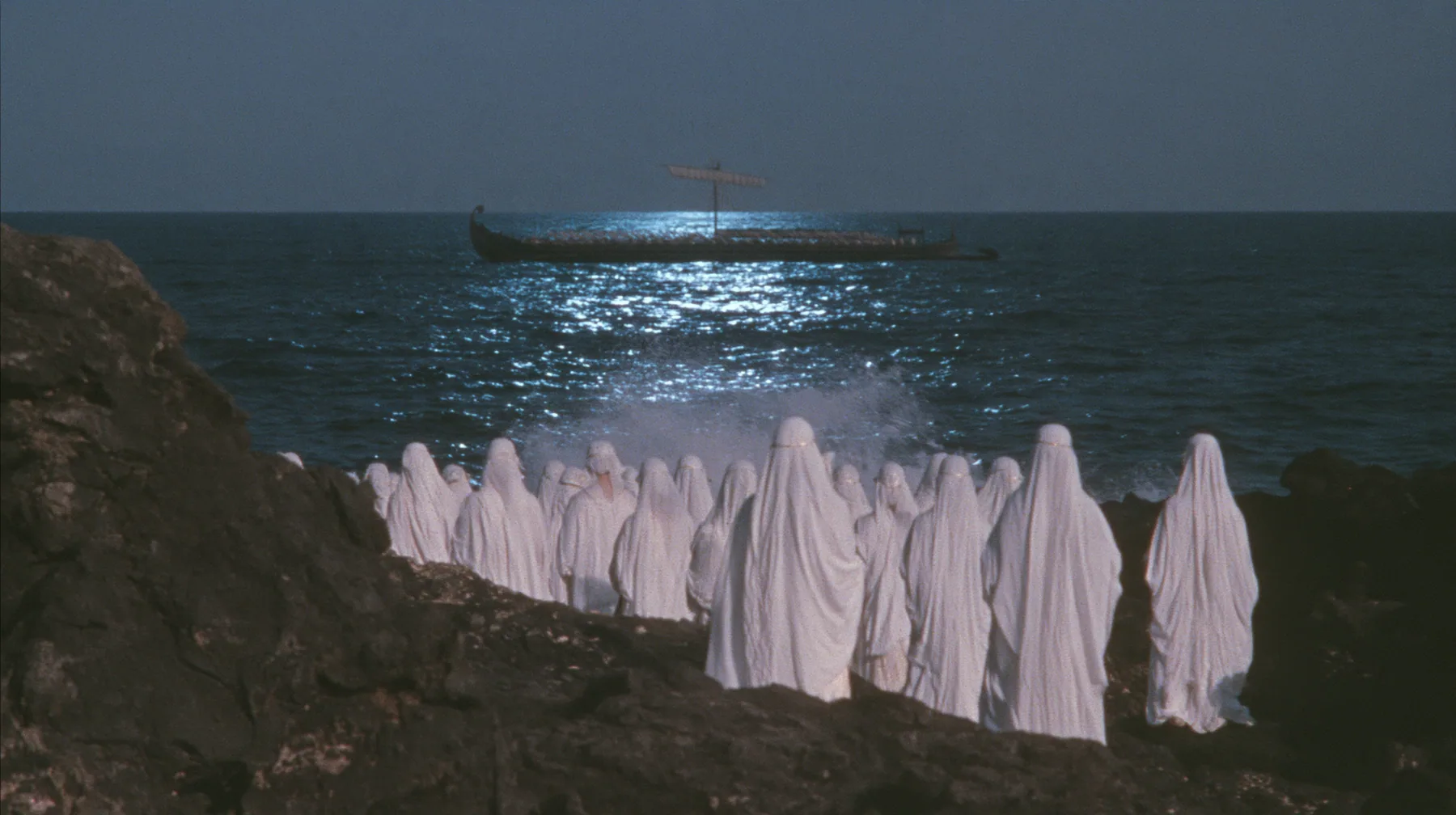 extreme wide shot from behind hundreds of women in white gowns on black sea rocks, looking out to a long black ancient Greek galley with a single mast, a square sail and two banks of oars on the far horizon, one…