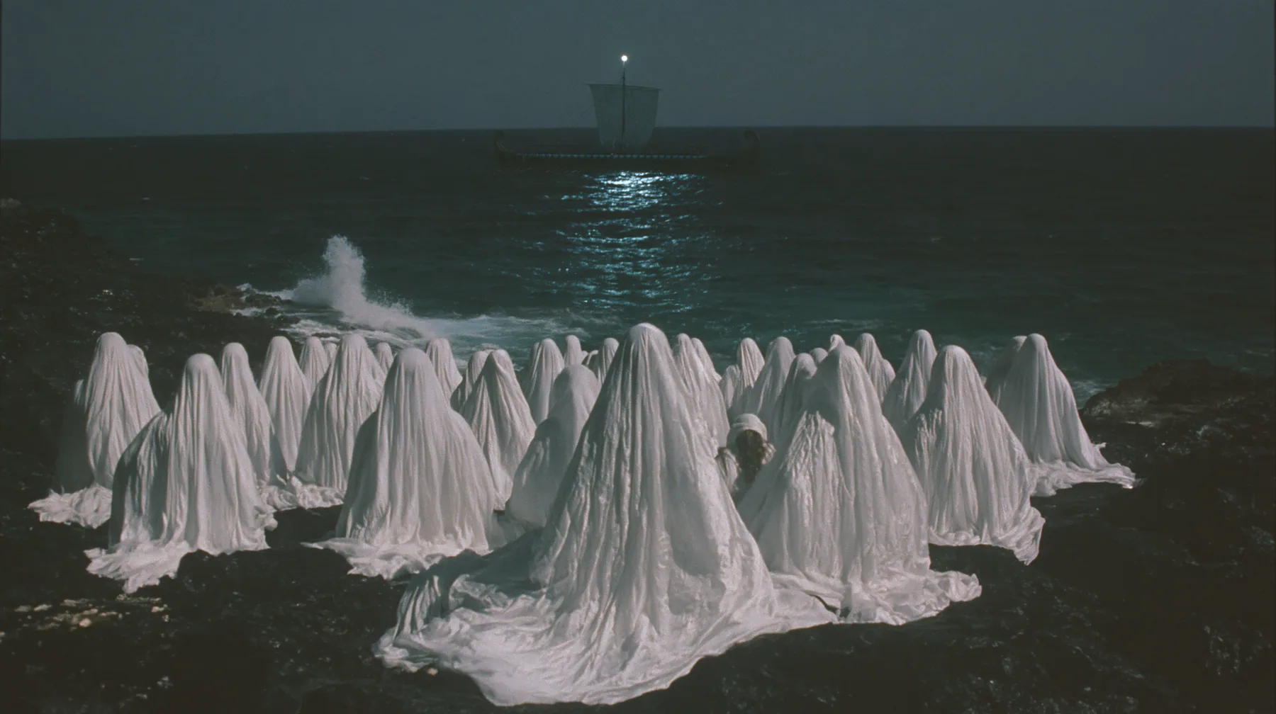 extreme wide shot from behind hundreds of women in white gowns on black sea rocks, looking out to a long black ancient Greek galley with a single mast, a square sail and two banks of oars on the far horizon, one…