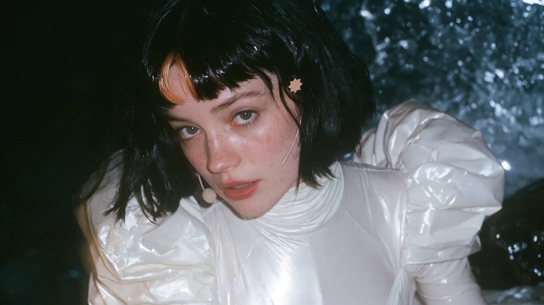 Claudia in CLODYSSEY: medium close-up of a Caucasian American woman in her mid-twenties with pale skin and light freckles, a glossy black blunt jaw-length bob with heavy straight bangs and one clay-orange streak through the bangs, a small…