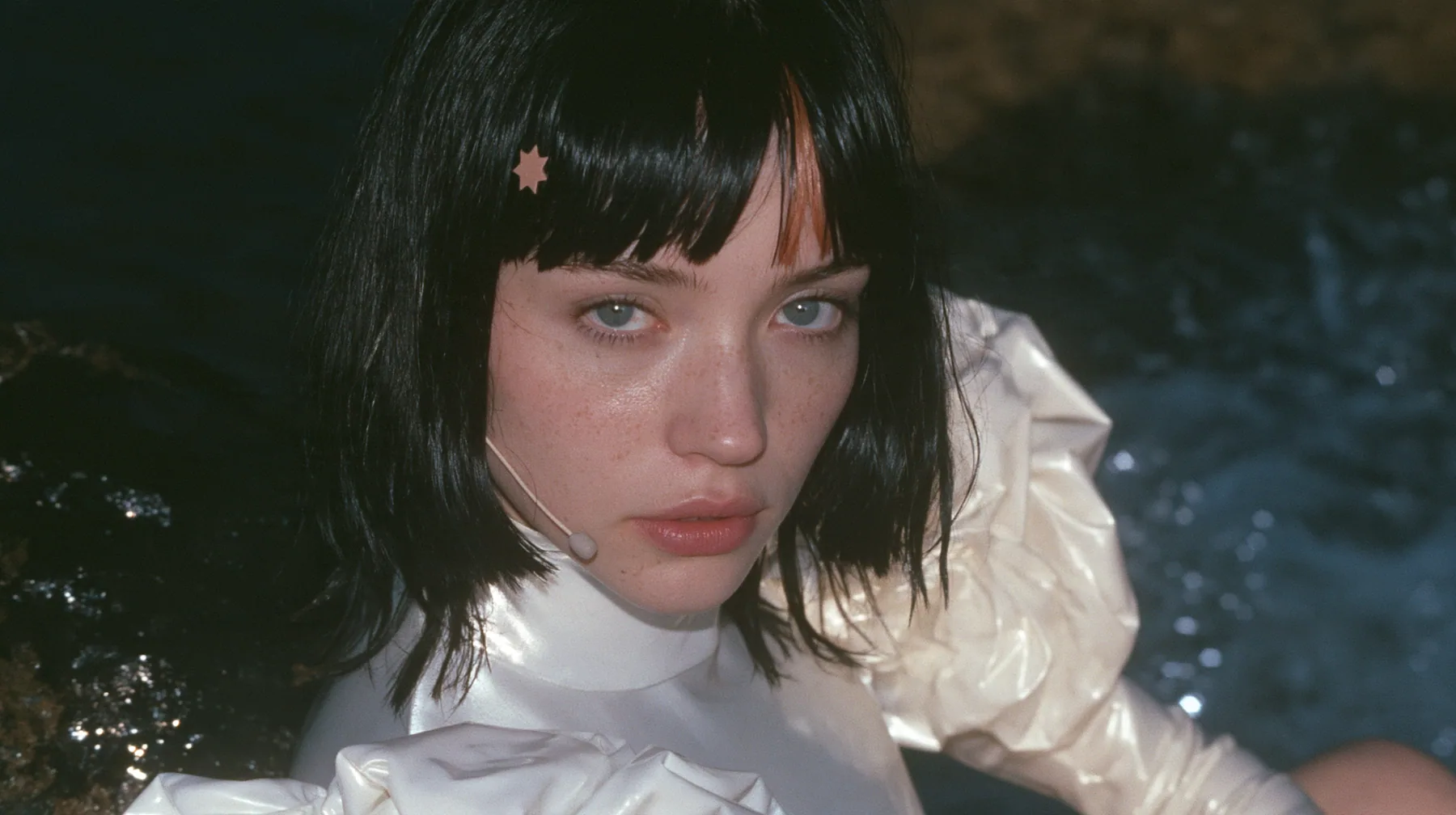 medium close-up of a Caucasian American woman in her mid-twenties with pale skin and light freckles, a glossy black blunt jaw-length bob with heavy straight bangs and one clay-orange streak through the bangs, a small…