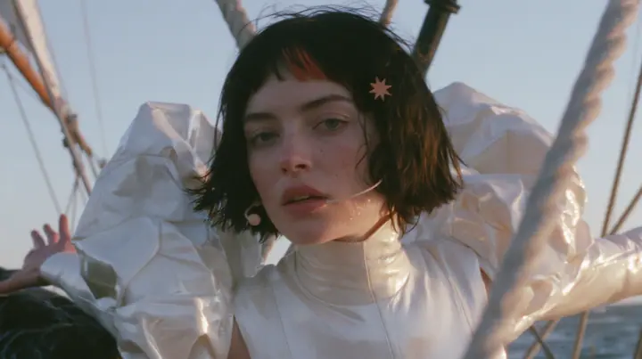 medium shot of a Caucasian American woman in her mid-twenties with pale skin and light freckles, a glossy black blunt jaw-length bob with heavy straight bangs and one clay-orange streak through the bangs, a small…