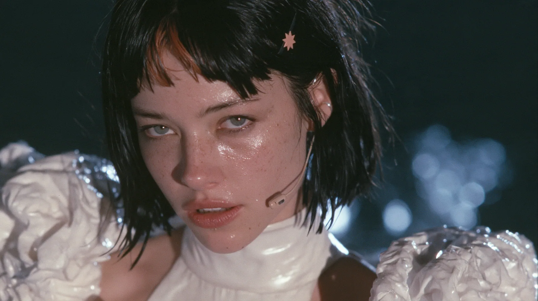 close-up of a Caucasian American woman in her mid-twenties with pale skin and light freckles, a glossy black blunt jaw-length bob with heavy straight bangs and one clay-orange streak through the bangs, a small flat…