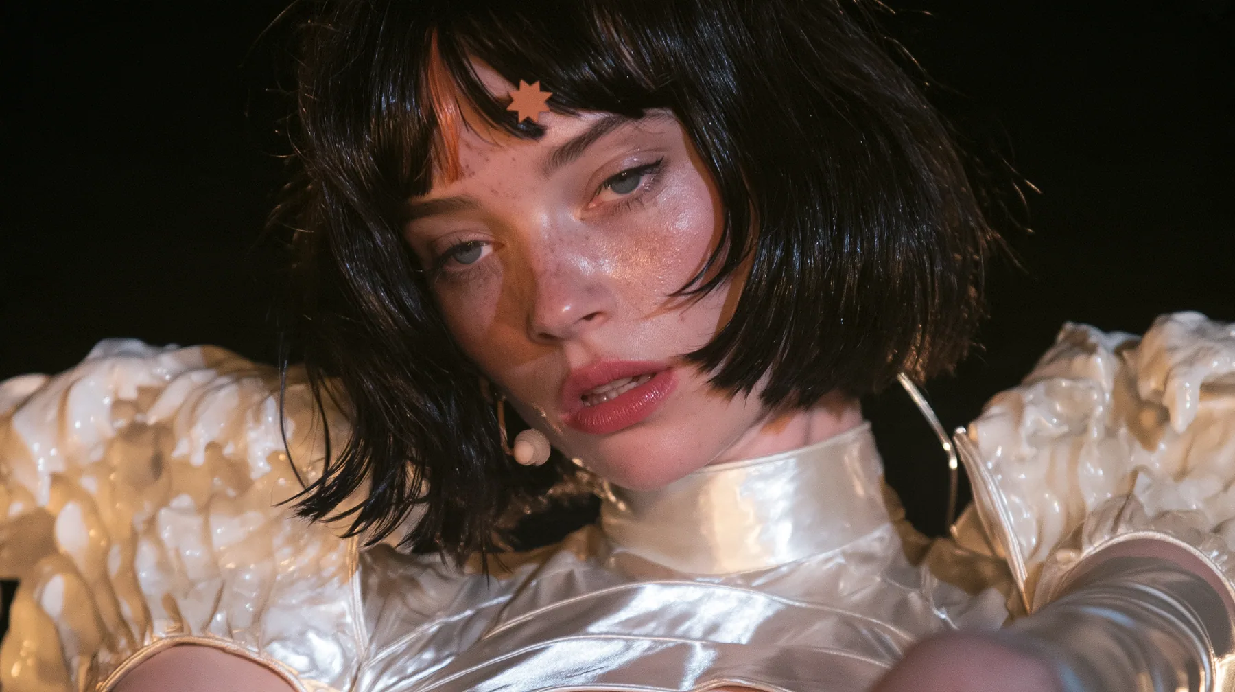 close-up of a Caucasian American woman in her mid-twenties with pale skin and light freckles, a glossy black blunt jaw-length bob with heavy straight bangs and one clay-orange streak through the bangs, a small flat…
