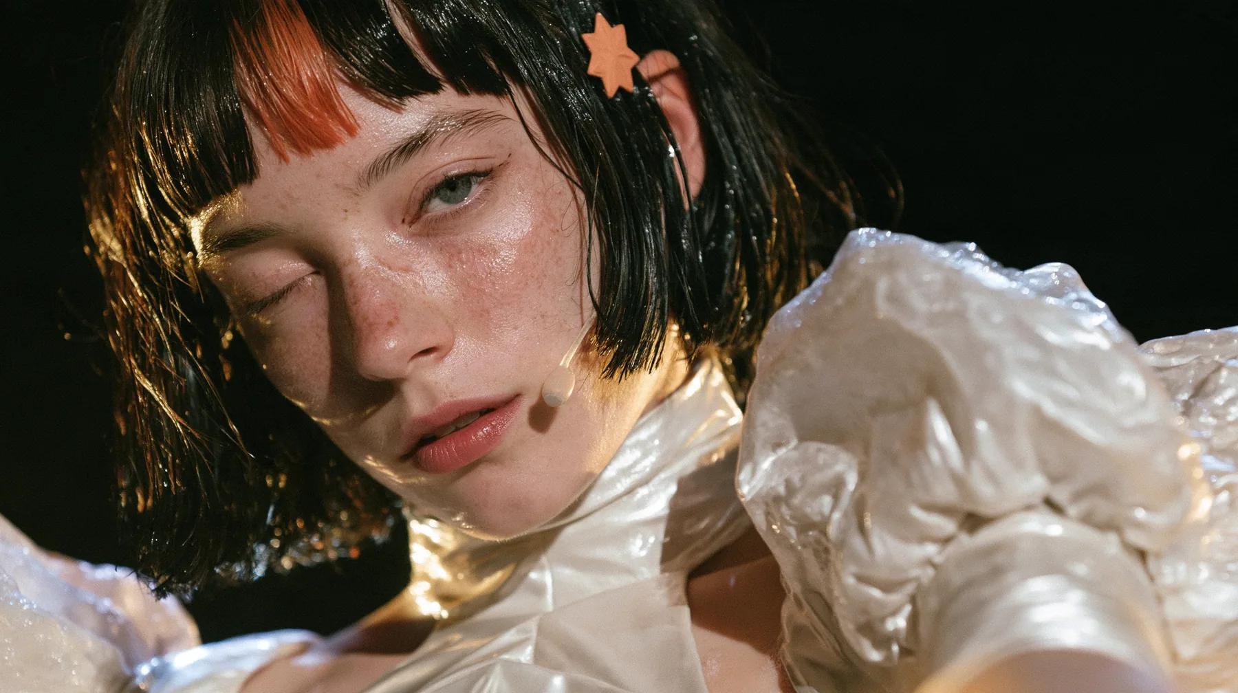 close-up of a Caucasian American woman in her mid-twenties with pale skin and light freckles, a glossy black blunt jaw-length bob with heavy straight bangs and one clay-orange streak through the bangs, a small flat…