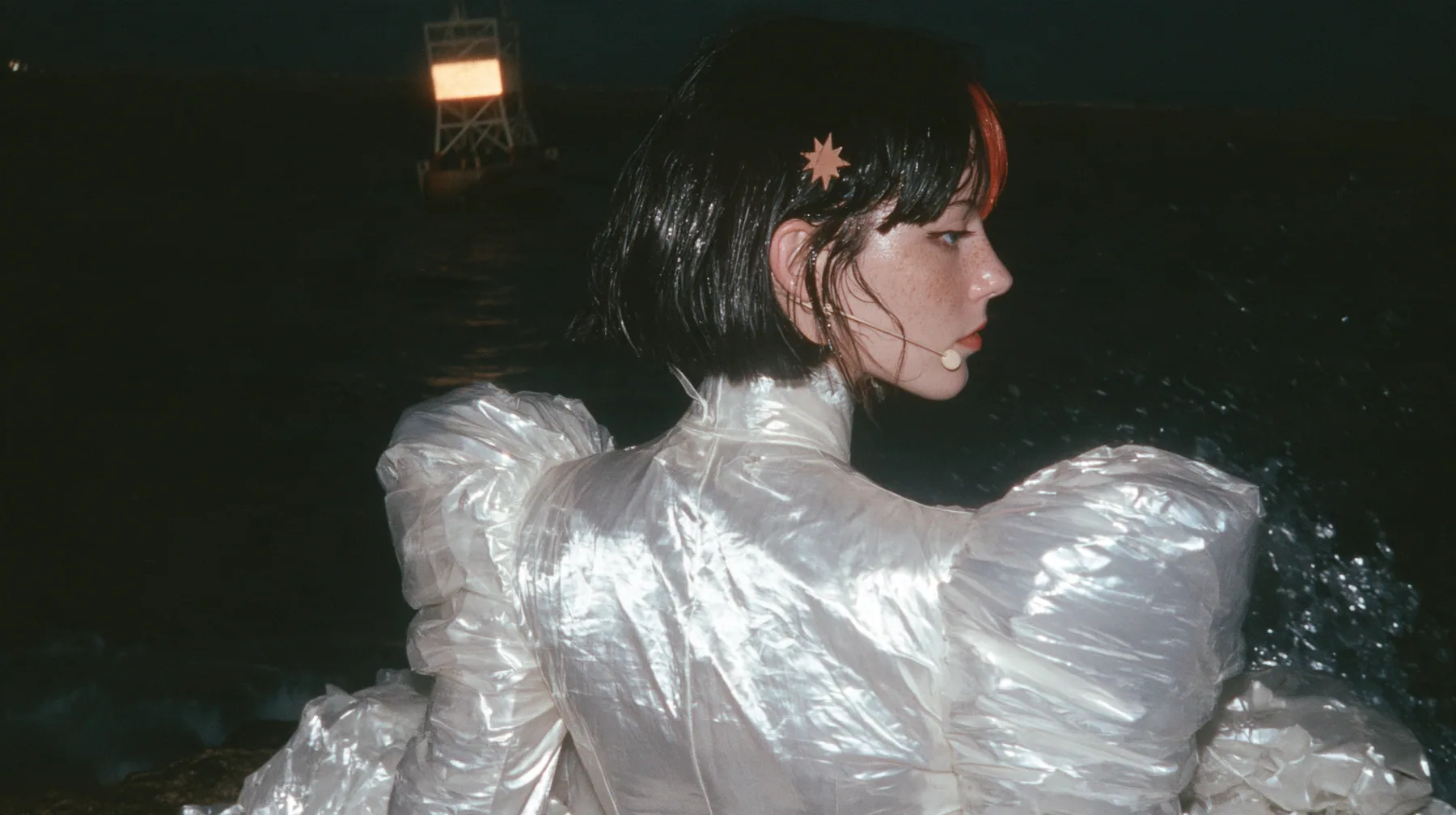over-the-shoulder shot from behind a Caucasian American woman in her mid-twenties with pale skin and light freckles, a glossy black blunt jaw-length bob with heavy straight bangs and one clay-orange streak through…