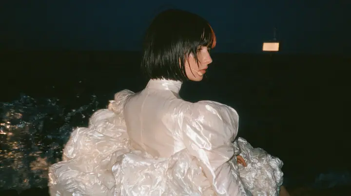 over-the-shoulder shot from behind a Caucasian American woman in her mid-twenties with pale skin and light freckles, a glossy black blunt jaw-length bob with heavy straight bangs and one clay-orange streak through…