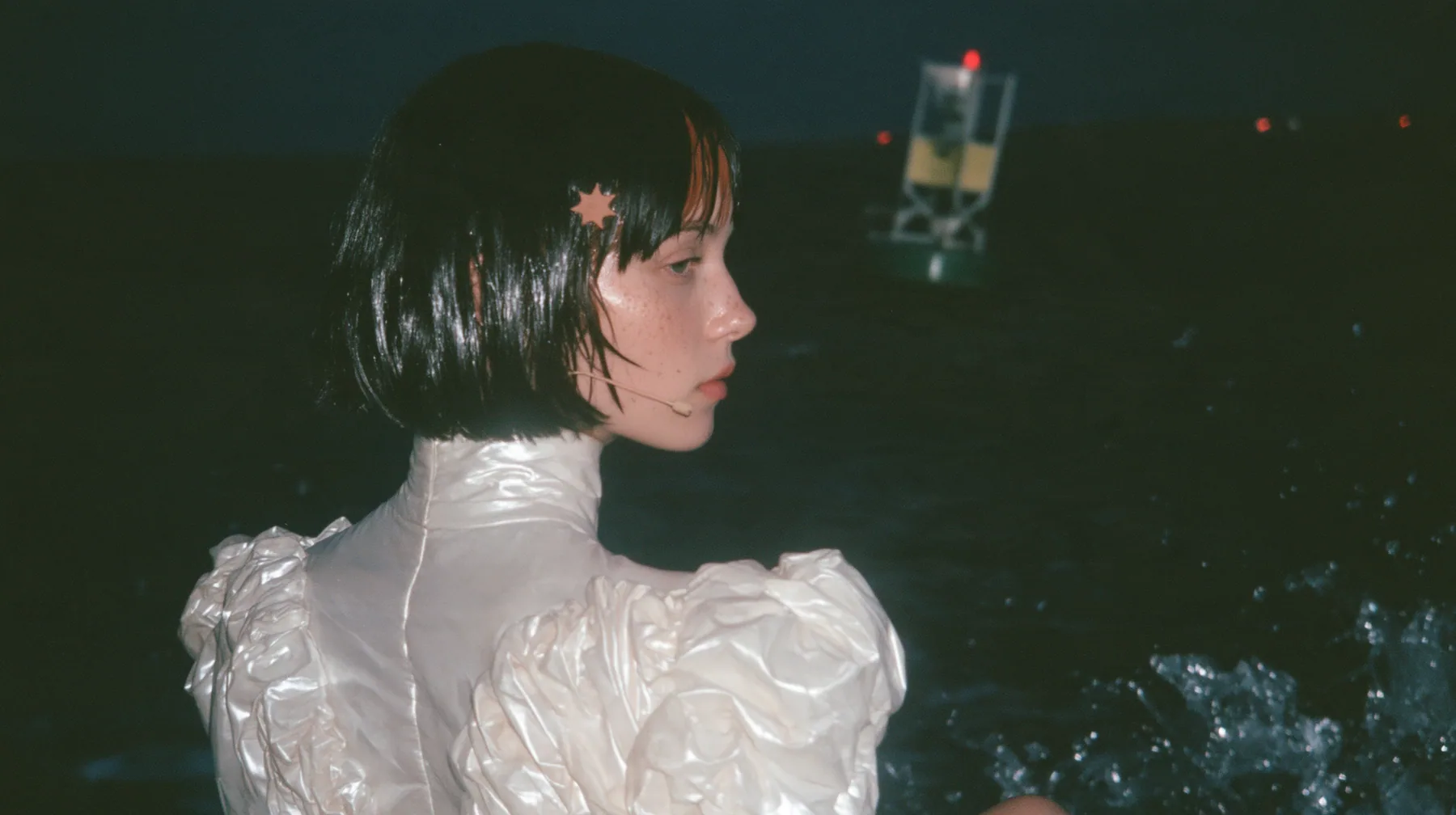 over-the-shoulder shot from behind a Caucasian American woman in her mid-twenties with pale skin and light freckles, a glossy black blunt jaw-length bob with heavy straight bangs and one clay-orange streak through…