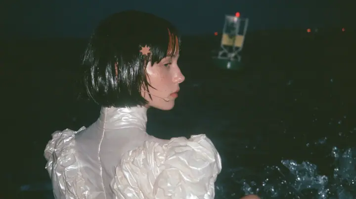 over-the-shoulder shot from behind a Caucasian American woman in her mid-twenties with pale skin and light freckles, a glossy black blunt jaw-length bob with heavy straight bangs and one clay-orange streak through…