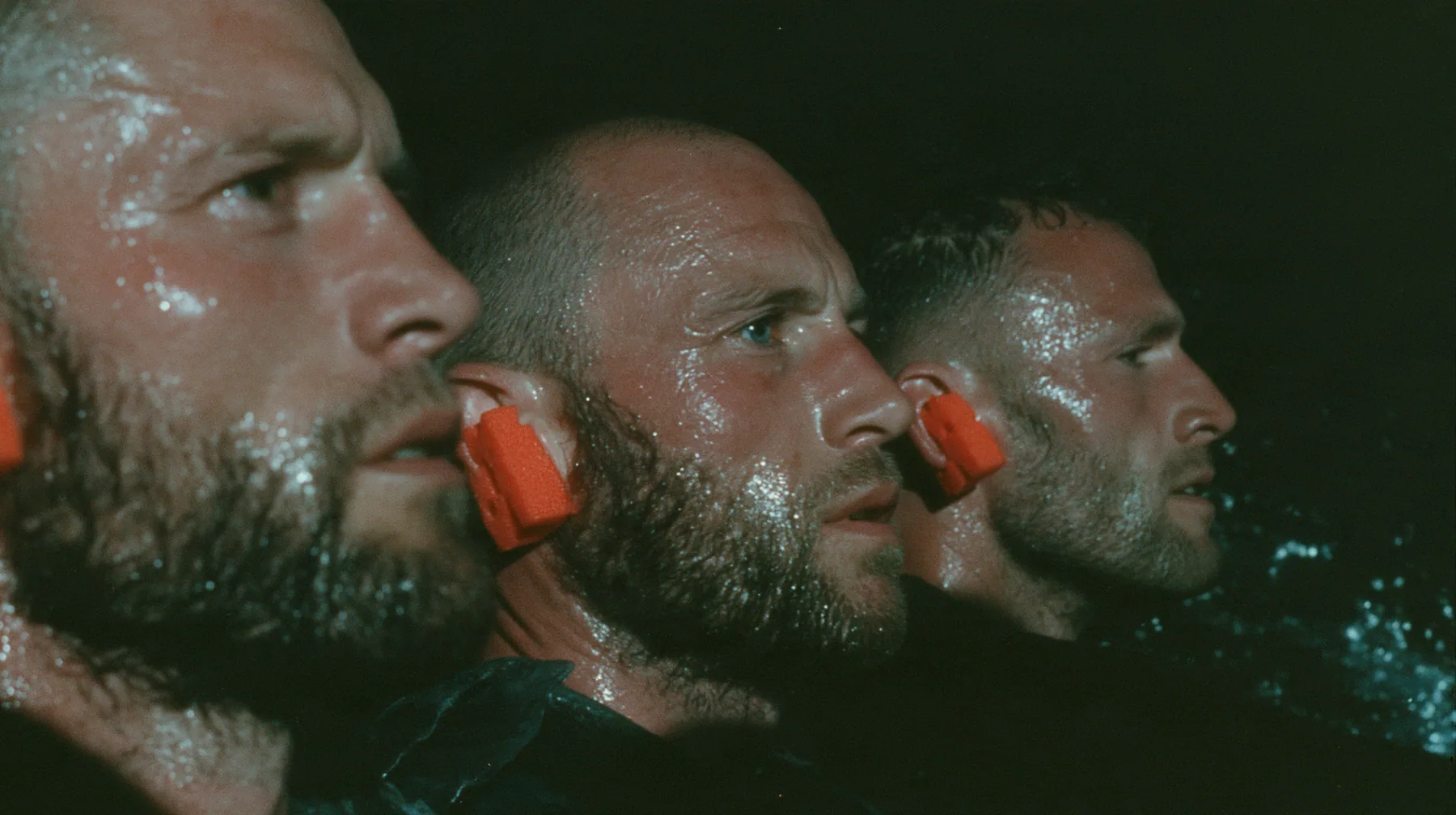close-up of the heads of three rowers in profile at the oars of a long black ancient Greek galley with a single mast, a square sail and two banks of oars at night, bright orange foam earplugs in their ears, moonlight…