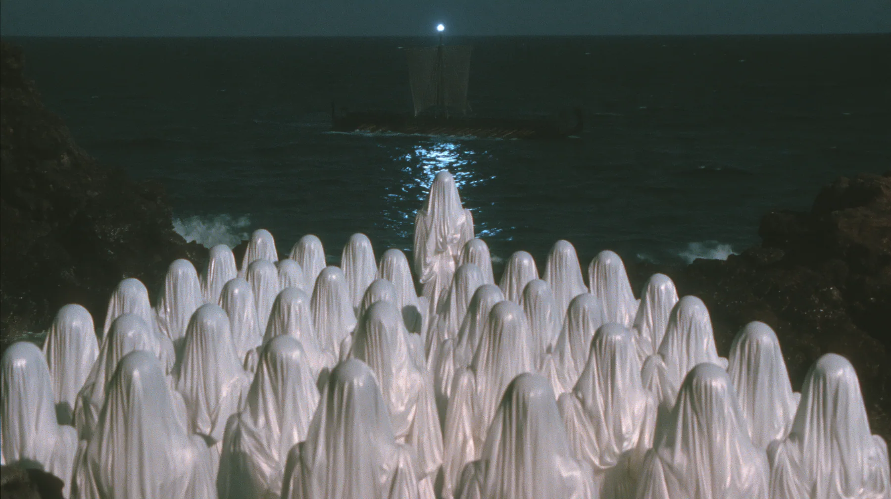 extreme wide shot from behind hundreds of identical bare-headed women with glossy black bobs in white couture gowns standing on black sea rocks with their backs to the camera, looking out to a long black ancient…