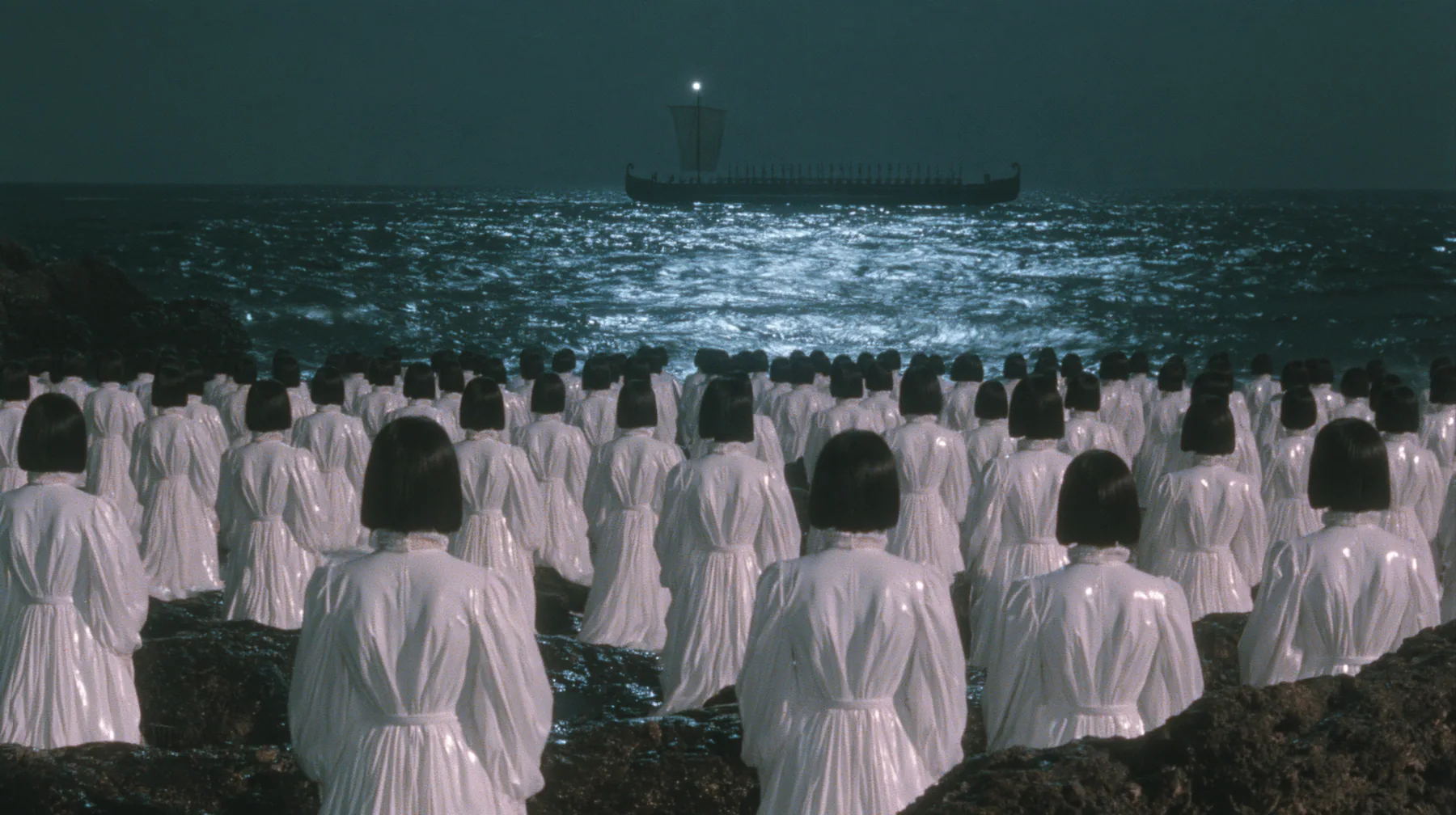 extreme wide shot from behind hundreds of identical bare-headed women with glossy black bobs in white couture gowns standing on black sea rocks with their backs to the camera, looking out to a long black ancient…