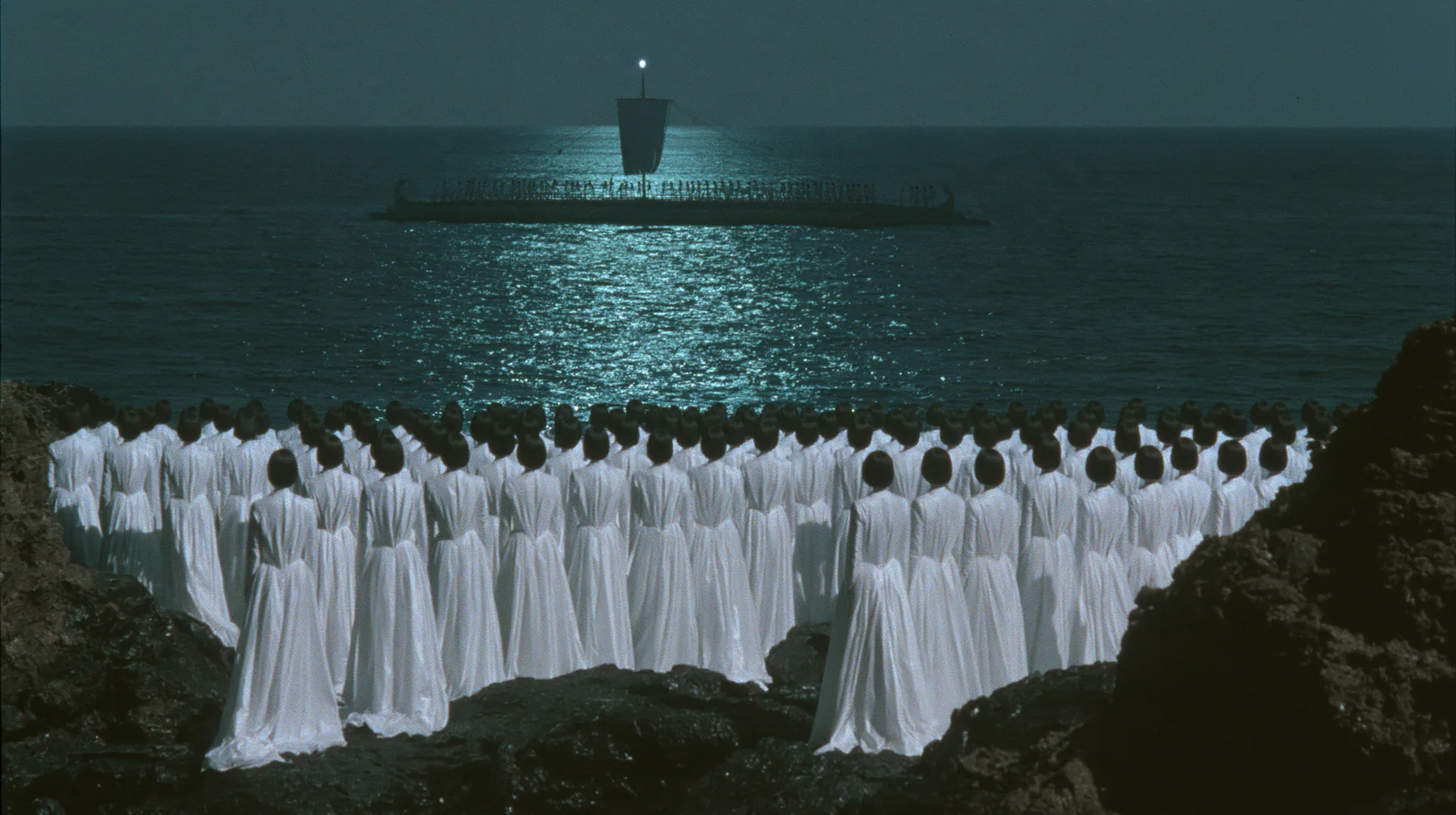 extreme wide shot from behind hundreds of identical bare-headed women with glossy black bobs in white couture gowns standing on black sea rocks with their backs to the camera, looking out to a long black ancient…