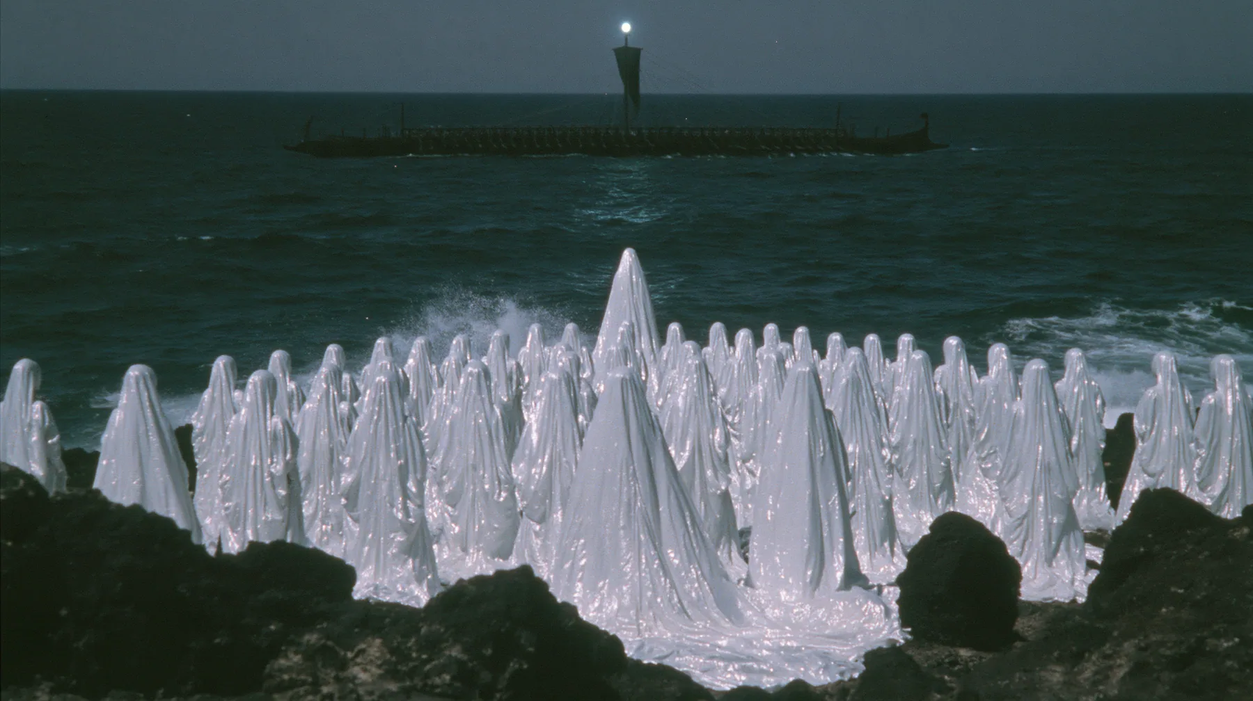 extreme wide shot from behind hundreds of identical bare-headed women with glossy black bobs in white couture gowns standing on black sea rocks with their backs to the camera, looking out to a long black ancient…