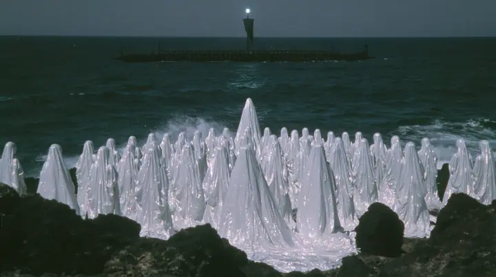 extreme wide shot from behind hundreds of identical bare-headed women with glossy black bobs in white couture gowns standing on black sea rocks with their backs to the camera, looking out to a long black ancient…