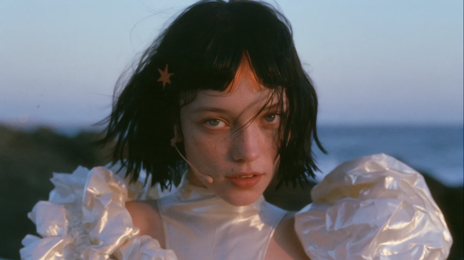 telephoto close-up of a Caucasian American woman in her mid-twenties with pale skin and light freckles, a glossy black blunt jaw-length bob with heavy straight bangs and one clay-orange streak through the bangs, a…
