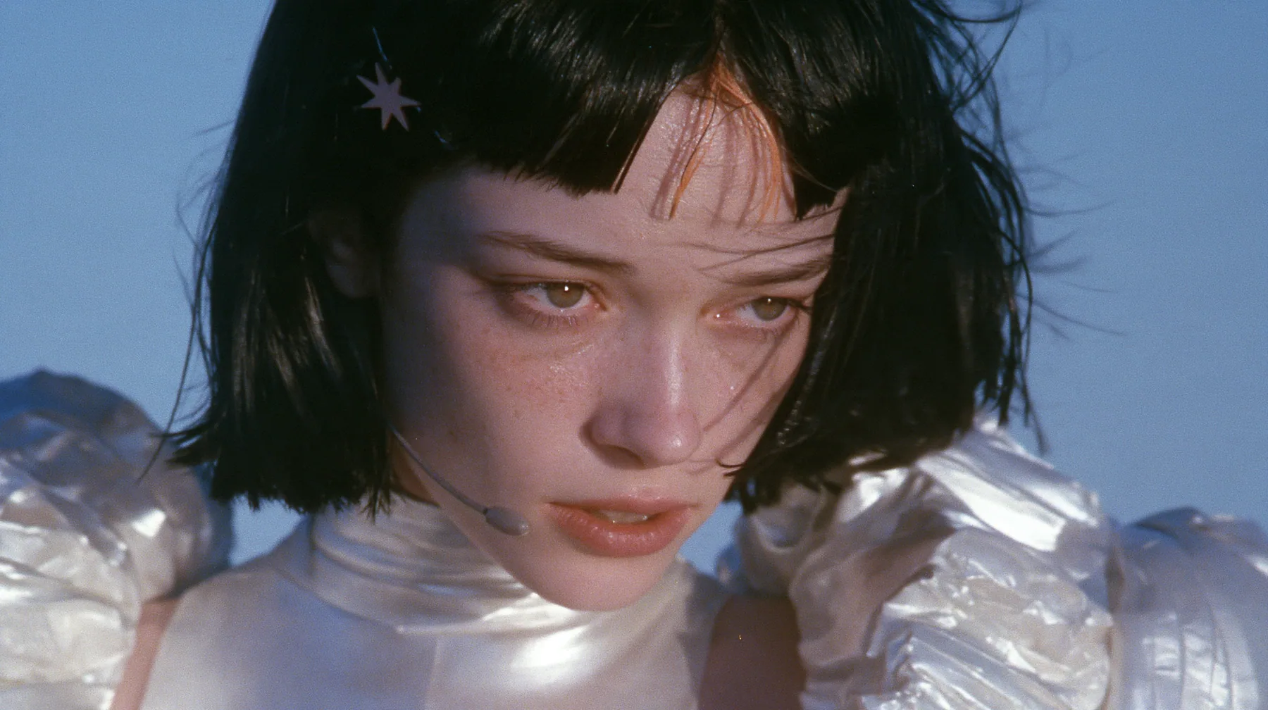 telephoto close-up of a Caucasian American woman in her mid-twenties with pale skin and light freckles, a glossy black blunt jaw-length bob with heavy straight bangs and one clay-orange streak through the bangs, a…