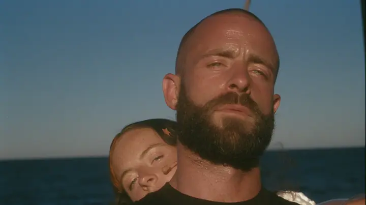 close-up of a broad-shouldered Caucasian man in his early thirties with a shaved head, a thick short dark beard, a square jaw, heavy brows and pale blue eyes, the build of a man who lifts, in a plain black t-shirt…