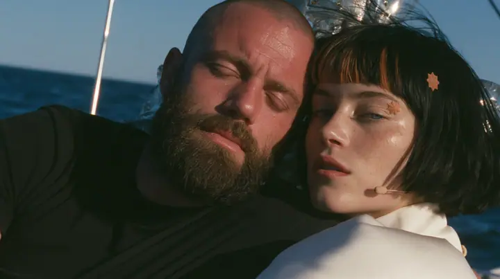 close-up of a broad-shouldered Caucasian man in his early thirties with a shaved head, a thick short dark beard, a square jaw, heavy brows and pale blue eyes, the build of a man who lifts, in a plain black t-shirt…