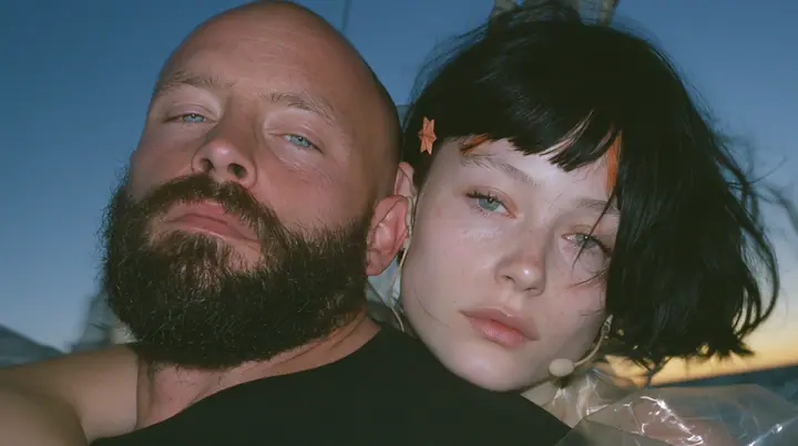 close-up of a broad-shouldered Caucasian man in his early thirties with a shaved head, a thick short dark beard, a square jaw, heavy brows and pale blue eyes, the build of a man who lifts, in a plain black t-shirt…