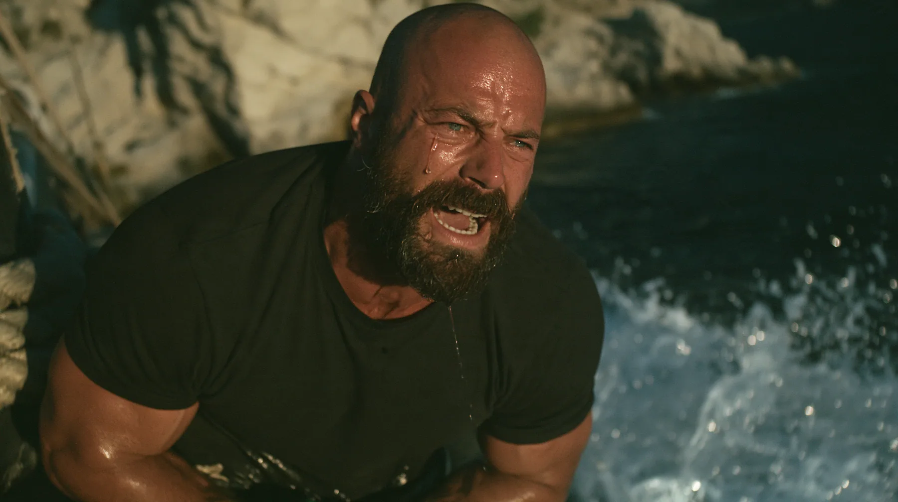 close-up of a broad-shouldered Caucasian man in his early thirties with a shaved head, a thick short dark beard, a square jaw, heavy brows and pale blue eyes, the build of a man who lifts, in a plain black t-shirt…