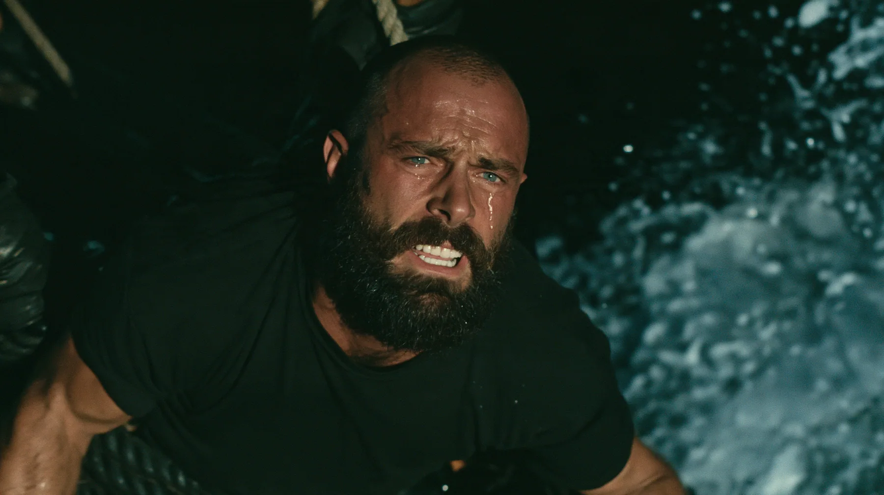 close-up of a broad-shouldered Caucasian man in his early thirties with a shaved head, a thick short dark beard, a square jaw, heavy brows and pale blue eyes, the build of a man who lifts, in a plain black t-shirt…