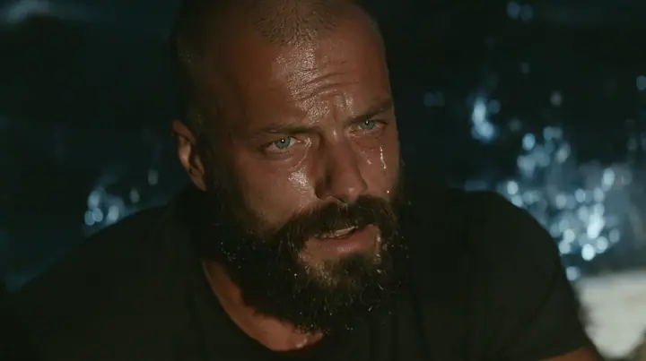 close-up of a broad-shouldered Caucasian man in his early thirties with a shaved head, a thick short dark beard, a square jaw, heavy brows and pale blue eyes, the build of a man who lifts, in a plain black t-shirt…