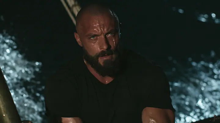 close-up of a broad-shouldered Caucasian man in his early thirties with a shaved head, a thick short dark beard, a square jaw, heavy brows and pale blue eyes, the build of a man who lifts, in a plain black t-shirt…