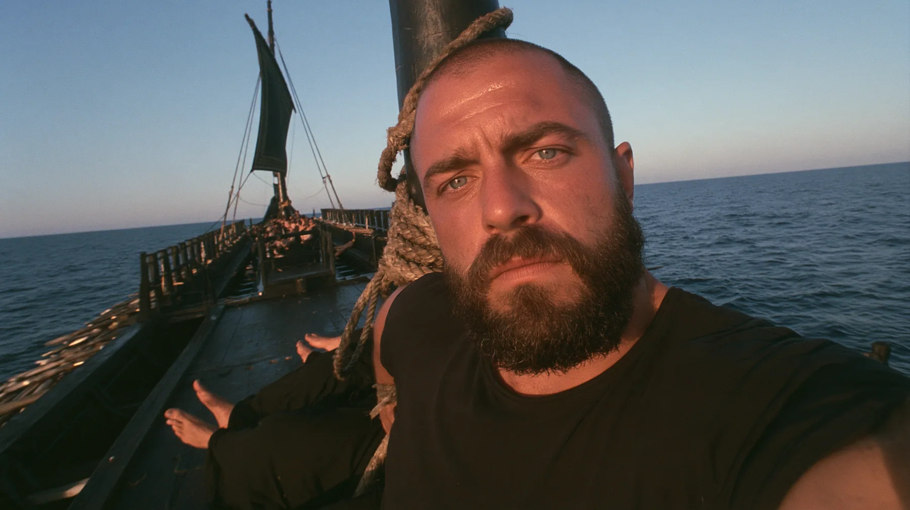 front-camera selfie view of a broad-shouldered Caucasian man in his early thirties with a shaved head, a thick short dark beard, a square jaw, heavy brows and pale blue eyes, the build of a man who lifts, in a plain…