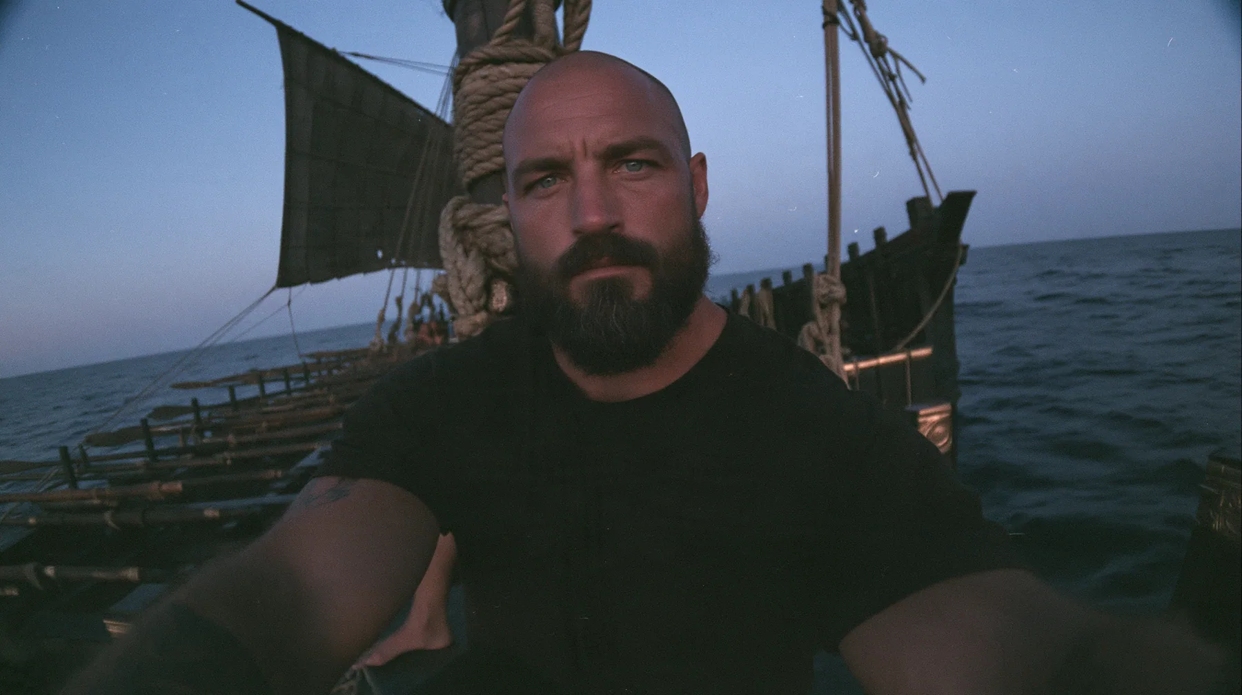 front-camera selfie view of a broad-shouldered Caucasian man in his early thirties with a shaved head, a thick short dark beard, a square jaw, heavy brows and pale blue eyes, the build of a man who lifts, in a plain…