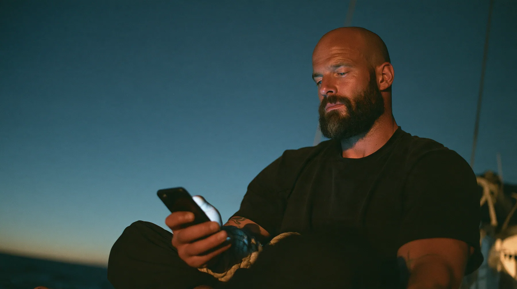 close-up of a broad-shouldered Caucasian man in his early thirties with a shaved head, a thick short dark beard, a square jaw, heavy brows and pale blue eyes, the build of a man who lifts, in a plain black t-shirt…