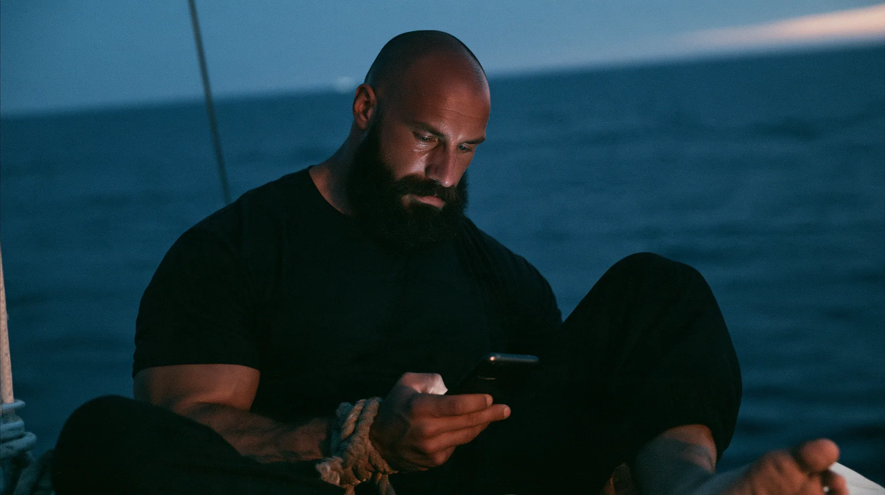 close-up of a broad-shouldered Caucasian man in his early thirties with a shaved head, a thick short dark beard, a square jaw, heavy brows and pale blue eyes, the build of a man who lifts, in a plain black t-shirt…