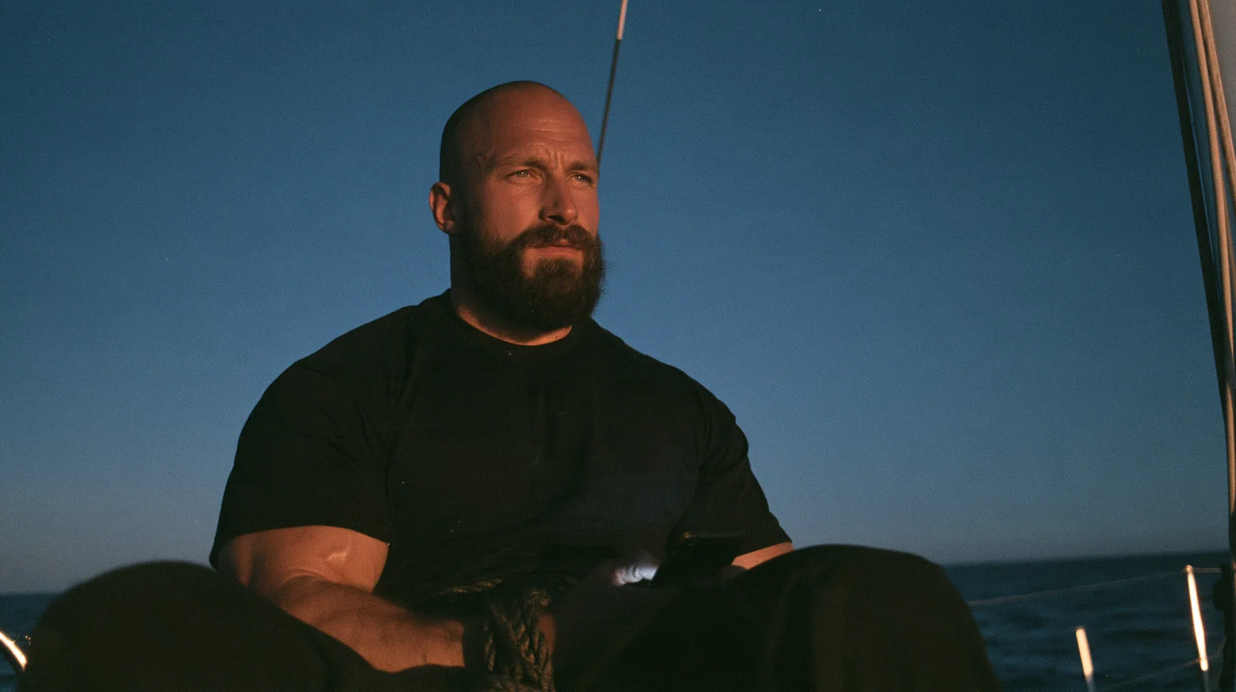 close-up of a broad-shouldered Caucasian man in his early thirties with a shaved head, a thick short dark beard, a square jaw, heavy brows and pale blue eyes, the build of a man who lifts, in a plain black t-shirt…