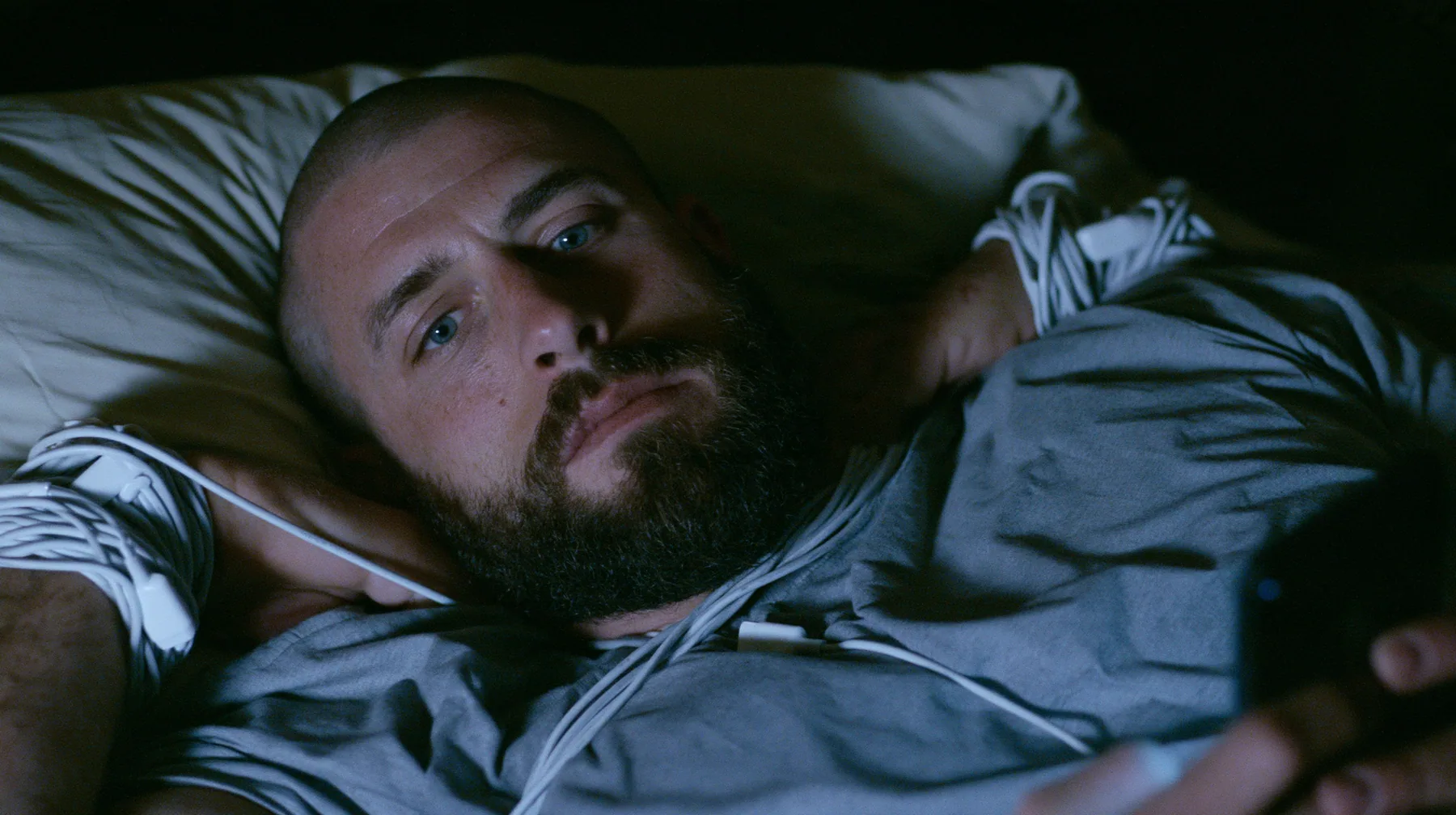 a broad-shouldered Caucasian man in his early thirties with a shaved head, a thick short dark beard, a square jaw, heavy brows and pale blue eyes, the build of a man who lifts, in a grey t-shirt, lying in bed with a…