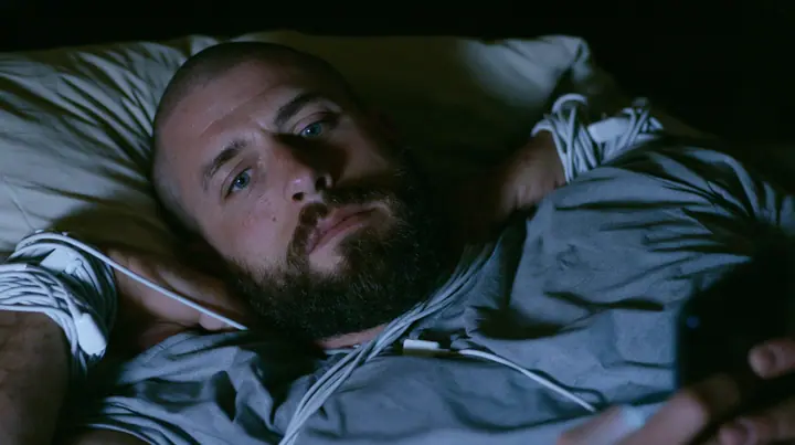 a broad-shouldered Caucasian man in his early thirties with a shaved head, a thick short dark beard, a square jaw, heavy brows and pale blue eyes, the build of a man who lifts, in a grey t-shirt, lying in bed with a…