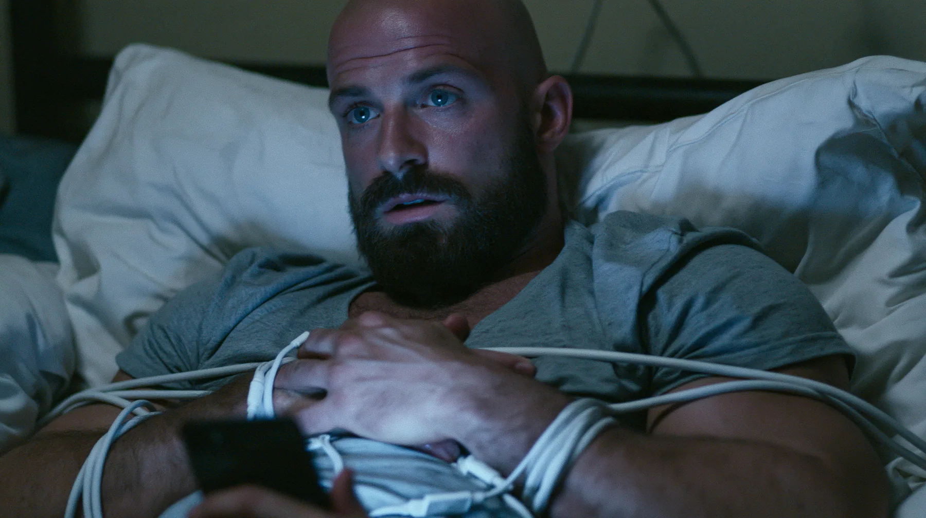 a broad-shouldered Caucasian man in his early thirties with a shaved head, a thick short dark beard, a square jaw, heavy brows and pale blue eyes, the build of a man who lifts, in a grey t-shirt, lying in bed with a…