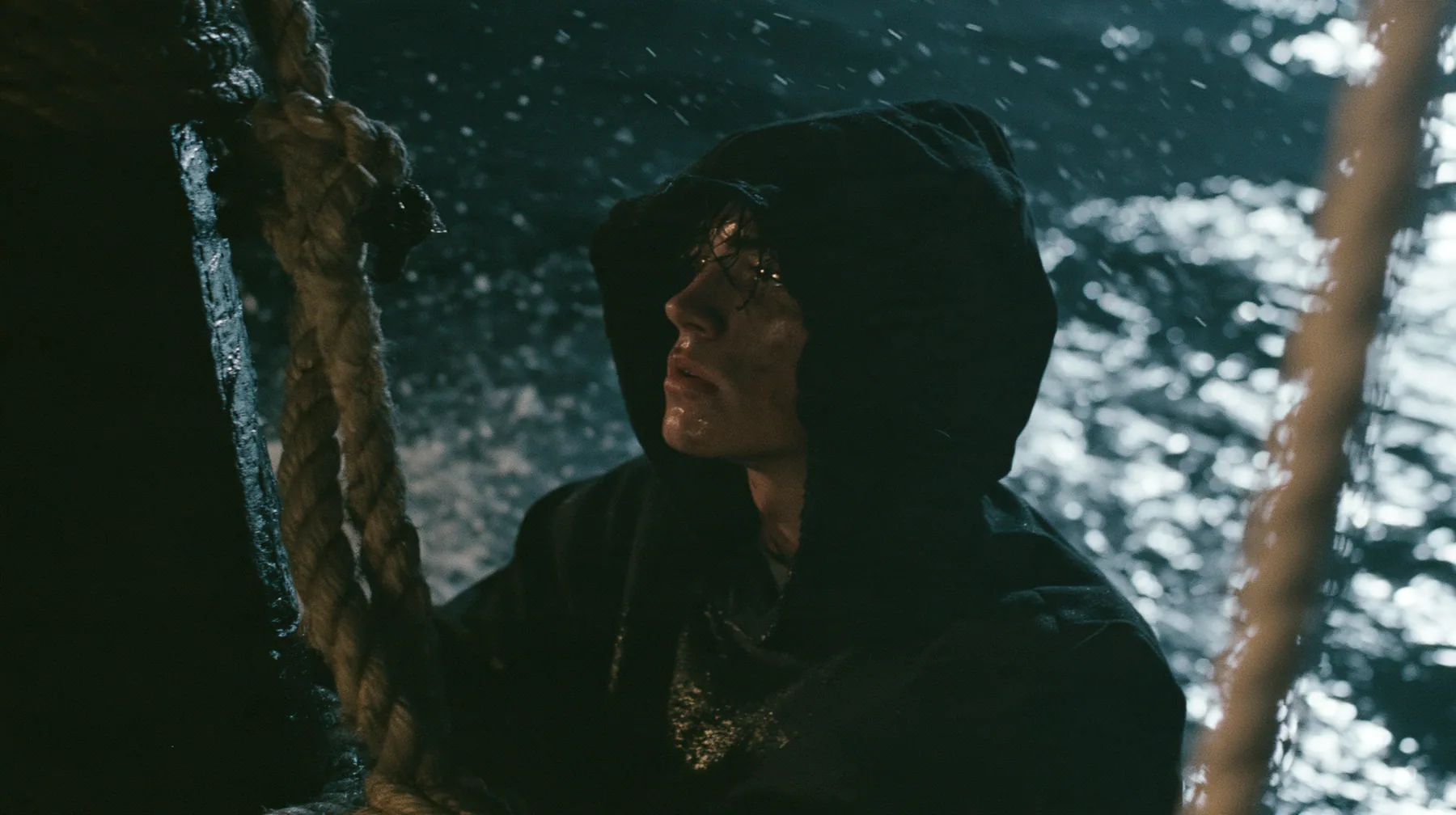 a gaunt young man in a black hoodie tied to the mast of a black ship with thick rope, night, moonlight on a black-teal sea, silver spray, cinematic 35mm film still, anamorphic, fine grain, no text, no letters, no logos