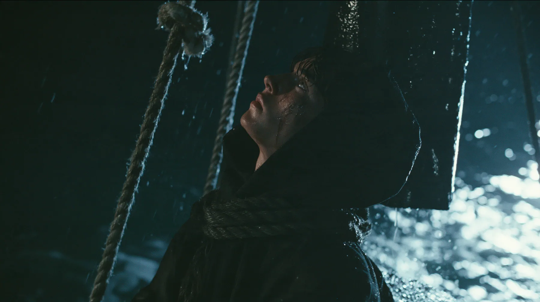 a gaunt young man in a black hoodie tied to the mast of a black ship with thick rope, night, moonlight on a black-teal sea, silver spray, cinematic 35mm film still, anamorphic, fine grain, no text, no letters, no logos