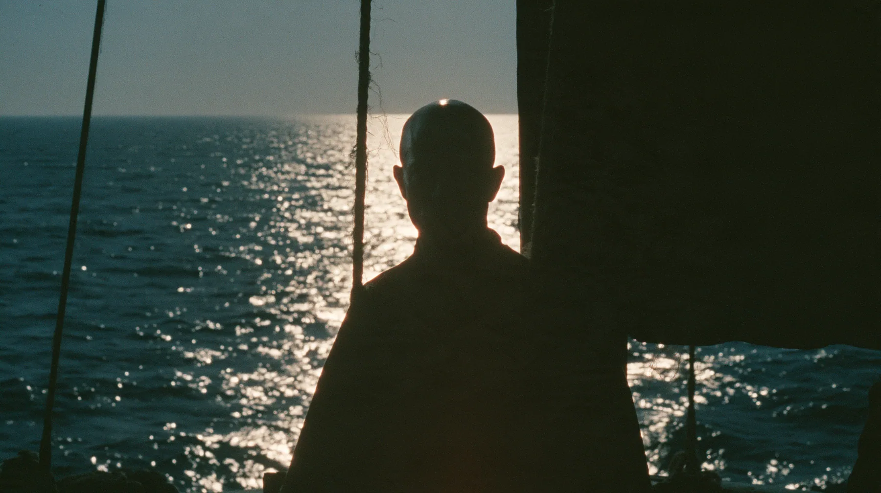 the silhouette of a bald man tied to the mast of a black ship against a lit sky, face in shadow, night, moonlight on a black-teal sea, silver spray, cinematic 35mm film still, anamorphic, fine grain, no text, no…