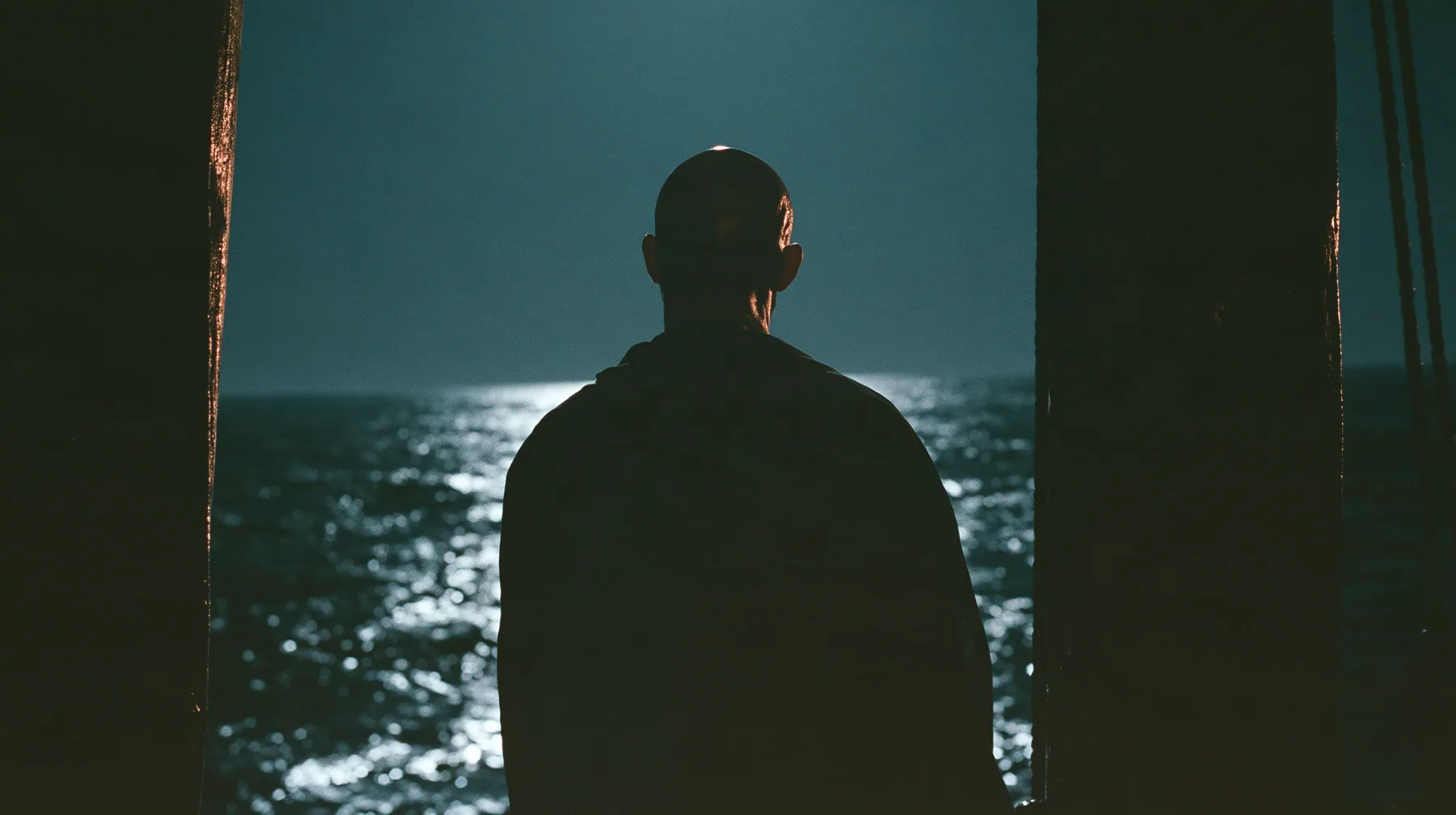 the silhouette of a bald man tied to the mast of a black ship against a lit sky, face in shadow, night, moonlight on a black-teal sea, silver spray, cinematic 35mm film still, anamorphic, fine grain, no text, no…