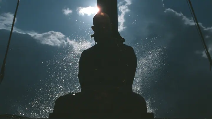 the silhouette of a bald man tied to the mast of a black ship against a lit sky, face in shadow, night, moonlight on a black-teal sea, silver spray, cinematic 35mm film still, anamorphic, fine grain, no text, no…